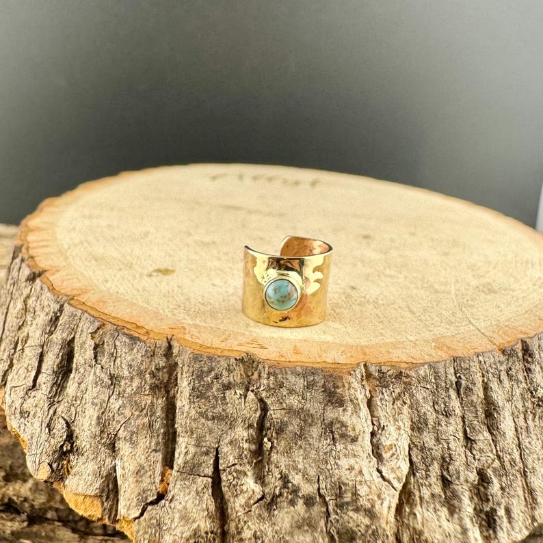 May include: A gold-colored ring with a turquoise-colored stone. The ring has a wide band with a textured surface and decorative cutouts. The turquoise stone is round and set in the center of the ring.