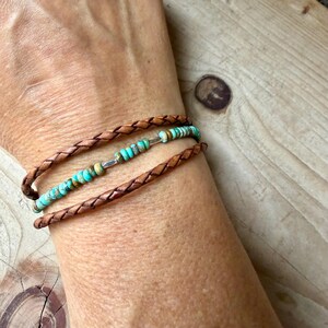 May include: A multi-strand bracelet featuring two braided brown leather cords and a central row of small, round, turquoise-colored beads with natural brown matrix patterns, accented by small silver-toned metal spacers between the beads.