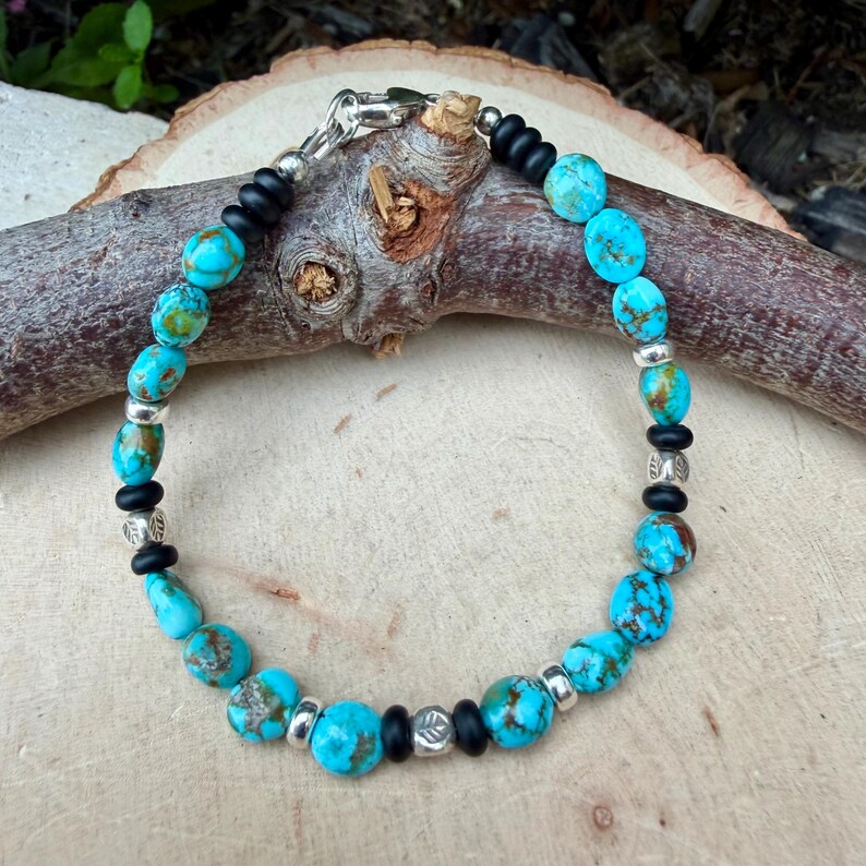 May include: A turquoise beaded bracelet with silver accents and black spacers. The bracelet features oval-shaped turquoise beads, interspersed with small silver beads and black rubber-like spacers. The clasp is silver.