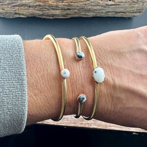 May include: Gold-toned metal bracelets with white and black speckled stone accents. The bracelets are open-ended, with the stones set at intervals along the bands. The jewelry is worn on a wrist, showcasing the design and craftsmanship.