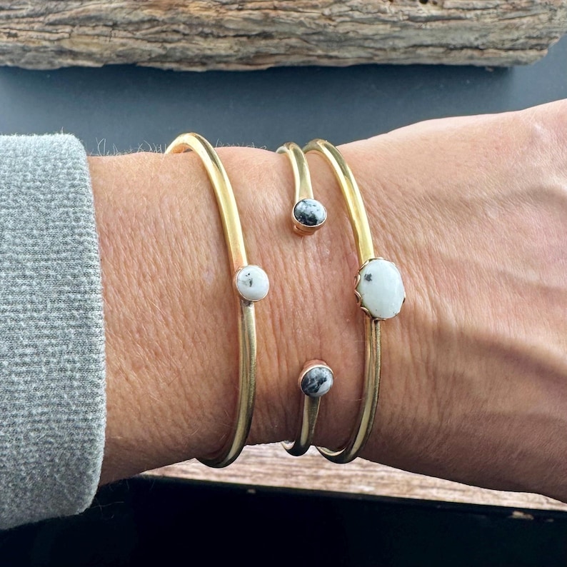 May include: Gold-toned metal bracelets with white and black speckled stone accents. The bracelets are open-ended, with the stones set at intervals along the bands. The jewelry is worn on a wrist, showcasing the design and craftsmanship.