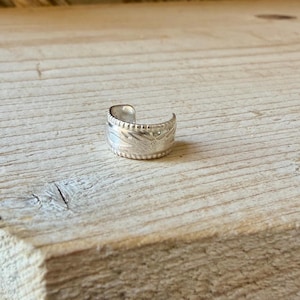 May include: A silver ear cuff featuring a wide band with a textured surface. The metal band has a repeating notched pattern along the top and bottom edges, with a central engraved design that wraps around the exterior of the circular piece.