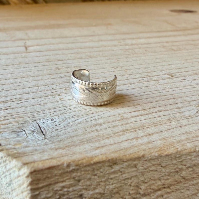 May include: A silver ear cuff featuring a wide band with a textured surface. The metal band has a repeating notched pattern along the top and bottom edges, with a central engraved design that wraps around the exterior of the circular piece.