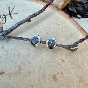 May include: Silver-coloured stud earrings in the shape of miniature cowboy hats. The earrings are oval with detailed hat brims and crowns. They are displayed on a wooden surface with a small branch.