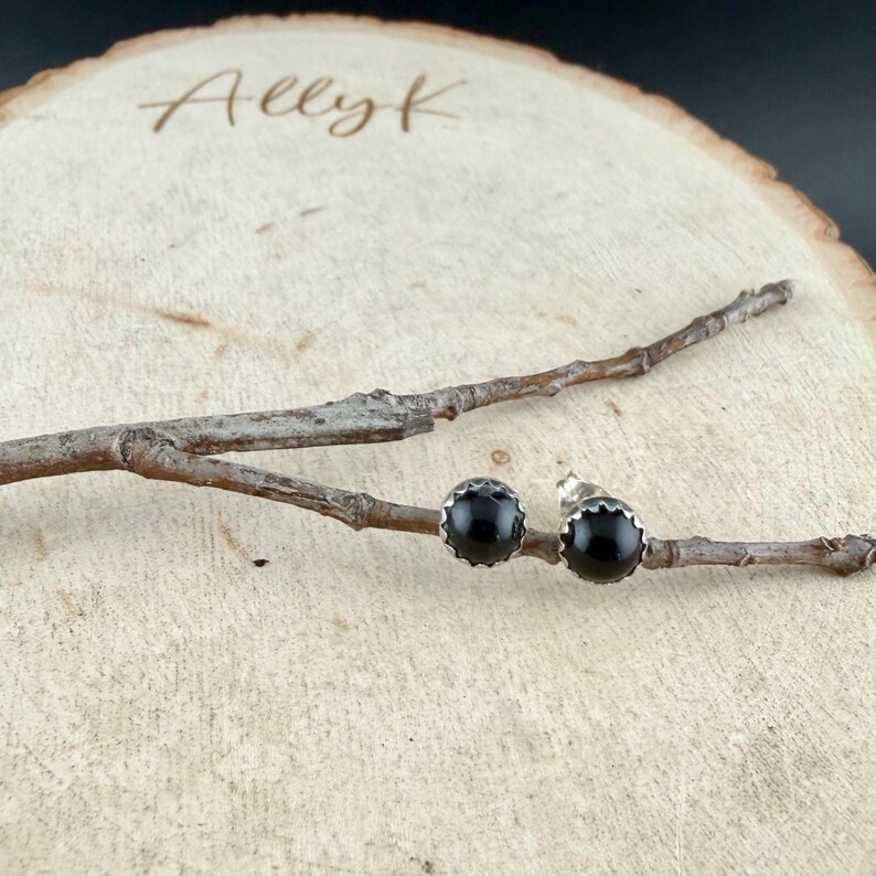 May include: A pair of black stud earrings with a scalloped silver setting. The earrings are displayed on a wooden surface with a small branch. The name "Ally K" is engraved on the wood.