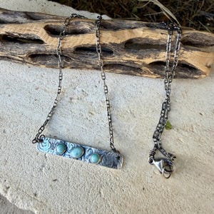 May include: A silver necklace featuring a rectangular bar pendant with an etched swirl pattern and three small, round, light blue stones set in the center. The pendant hangs from a dark, oxidized silver chain with a lobster claw clasp.
