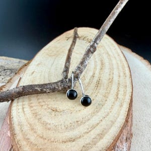 May include: A pair of small, round black onyx earrings with silver-colored hooks. The earrings are displayed on a wooden branch, set against a light wood grain background. The earrings are simple and elegant.