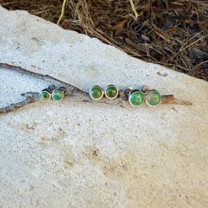 May include: Three pairs of stud earrings featuring round, polished green stones set in silver-toned metal settings. Each earring has a smooth, cabochon-style surface with natural color variations, displayed in a row on a thin, textured twig.