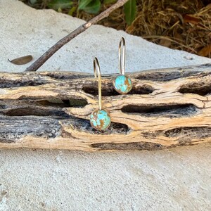 May include: A pair of drop earrings featuring round, polished turquoise stones with brown matrix patterns. Each stone is set in a simple gold-toned metal bezel and attached to a slender, curved gold-toned wire hook for hanging from the earlobe.