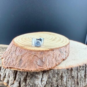 K&ouml;nnte beinhalten: Ein silberfarbener Ring mit breitem Band und einem kreisf&ouml;rmigen, hellblauen Stein. Der Ring befindet sich auf einer rustikalen Holzoberfl&auml;che, die sein Design und seine Textur hervorhebt. Das Band des Rings hat ein strukturiertes Muster.