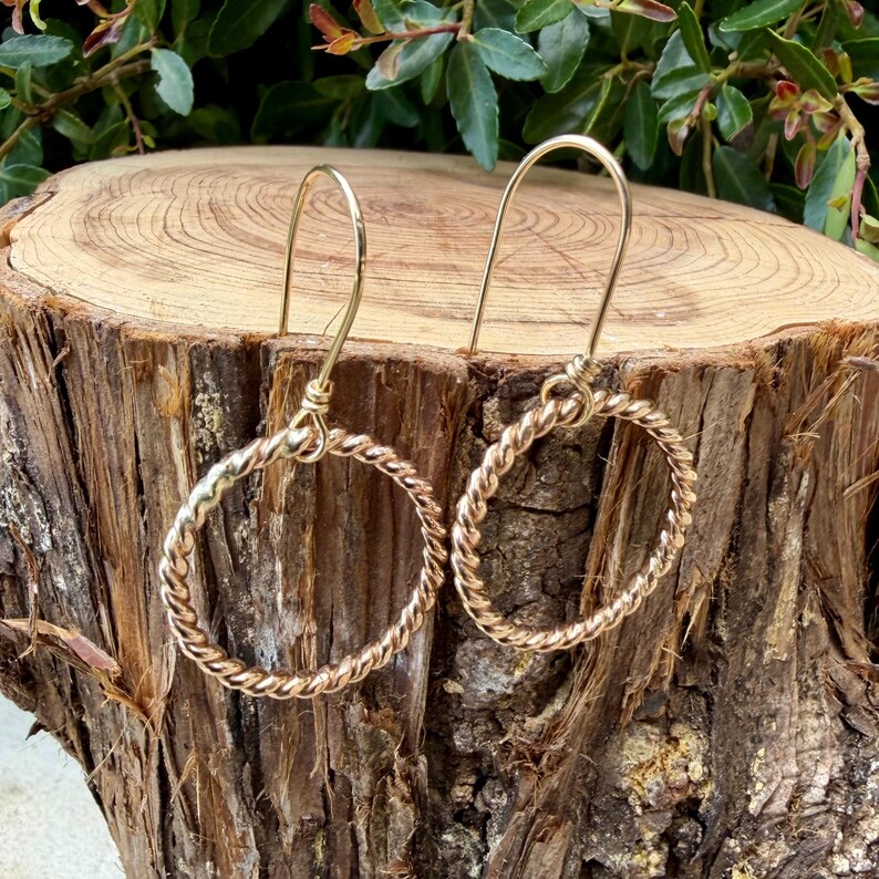 May include: A pair of gold-toned earrings featuring a circular hoop design with a twisted, rope-like texture. Each earring is suspended from a simple, curved wire hook that attaches to the top of the circular hoop, creating a dangling silhouette.