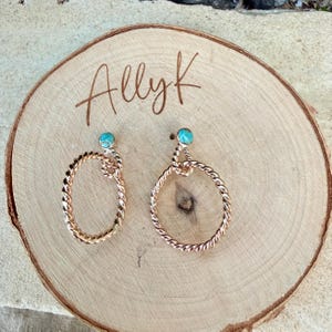 May include: A pair of rose gold earrings with a twisted rope design. Each earring features a small, round, turquoise-colored stone at the top. The earrings are displayed on a wooden surface with the handwritten text "Allyk".