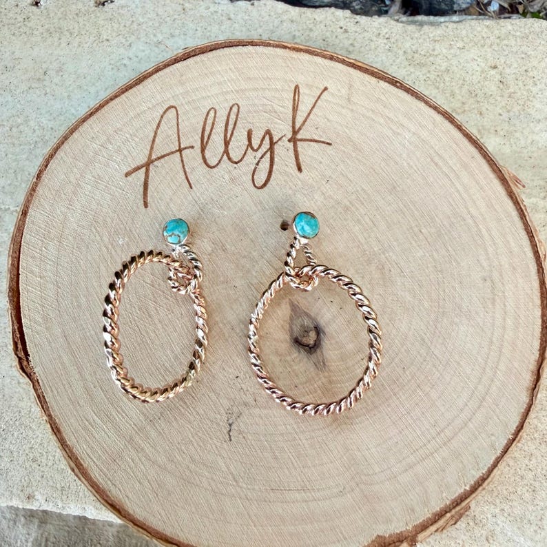 May include: A pair of rose gold earrings with a twisted rope design. Each earring features a small, round, turquoise-colored stone at the top. The earrings are displayed on a wooden surface with the handwritten text "Allyk".