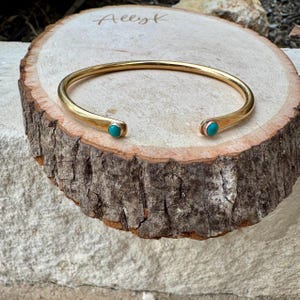 May include: A gold-colored cuff bracelet with turquoise-colored stones set at each end. The bracelet is displayed on a rustic, circular wooden surface. The wood has a natural, rough texture. The word "Allyk" is written on the wood.