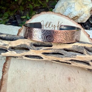 Puede incluir: Un brazalete de cobre con un intrincado dise&ntilde;o floral en espiral. La banda de metal es ancha y tiene un interior oscuro. El brazalete se exhibe sobre un trozo de madera. El texto "Helly K" es visible.