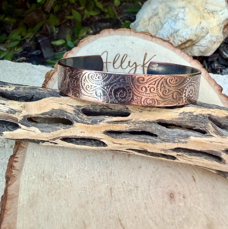 Puede incluir: Un brazalete de cobre con un intrincado dise&ntilde;o floral en espiral. La banda de metal es ancha y tiene un interior oscuro. El brazalete se exhibe sobre un trozo de madera. El texto "Helly K" es visible.