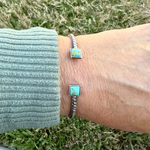 Op de afbeelding: Een zilverkleurige manchetarmband met een gedraaid touwontwerp. De armband heeft twee vierkante turkooizen stenen gezet in zilver. De armband is open, waardoor hij gemakkelijk aan en uit te trekken is.