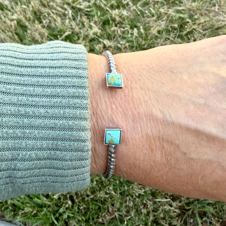 Op de afbeelding: Een zilverkleurige manchetarmband met een gedraaid touwontwerp. De armband heeft twee vierkante turkooizen stenen gezet in zilver. De armband is open, waardoor hij gemakkelijk aan en uit te trekken is.
