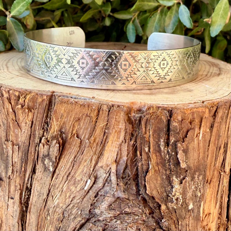 May include: A silver cuff bracelet featuring a repeating geometric pattern of triangles and diamonds engraved across the outer surface. The interior of the metal band is clearly stamped with the number 925, indicating the silver content of the piece.