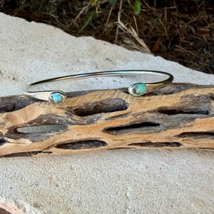May include: A silver cuff bracelet featuring an open design with two polished ends. Each end is set with a small, oval-shaped turquoise stone displaying natural blue and green colour variations and dark matrix veining throughout.