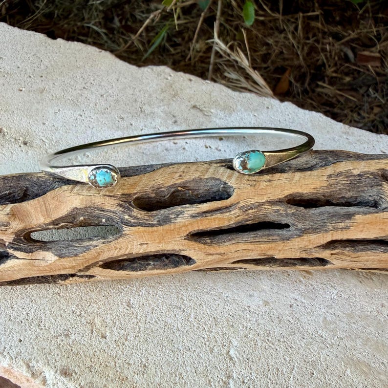 May include: A silver cuff bracelet featuring an open design with two polished ends. Each end is set with a small, oval-shaped turquoise stone displaying natural blue and green colour variations and dark matrix veining throughout.