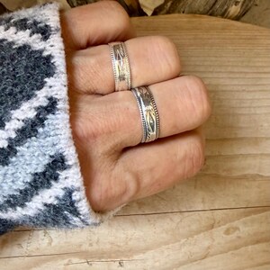 May include: Two silver rings featuring intricate engraved patterns and beaded edges. The rings are worn on adjacent fingers, showcasing a metallic finish with dark oxidized detailing within the etched designs on the bands.