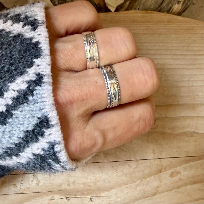 May include: Two silver rings featuring intricate engraved patterns and beaded edges. The rings are worn on adjacent fingers, showcasing a metallic finish with dark oxidized detailing within the etched designs on the bands.