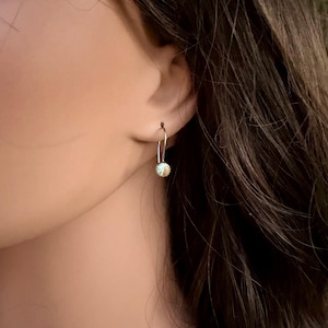 May include: A silver-toned earring featuring a delicate curved wire hook that suspends a small, spherical charm. The charm has a textured, glittery surface with a light blue and gold color palette, reflecting light with a subtle shimmer.