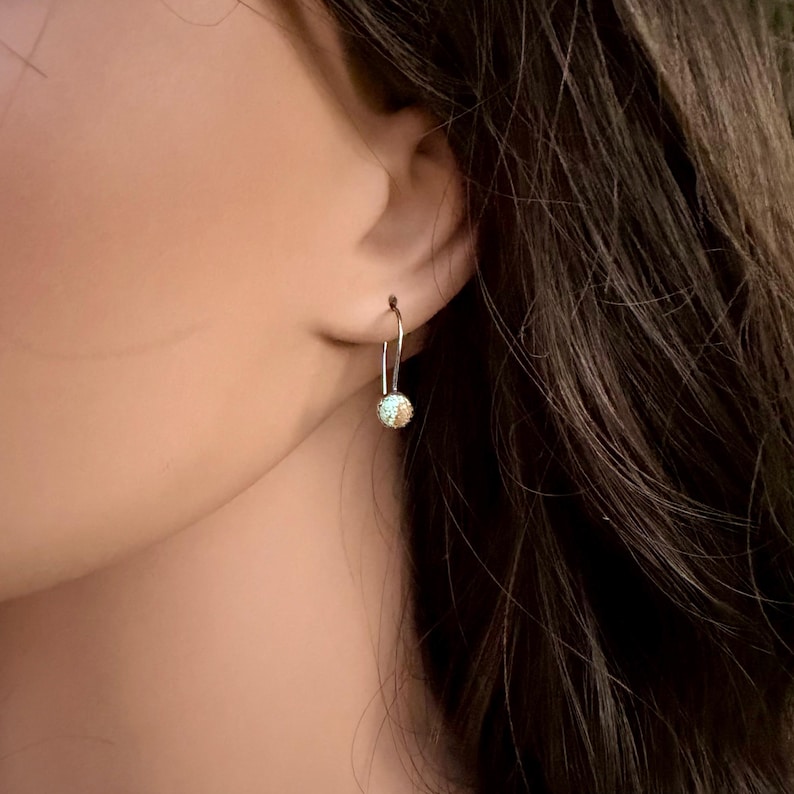 May include: A silver-toned earring featuring a delicate curved wire hook that suspends a small, spherical charm. The charm has a textured, glittery surface with a light blue and gold color palette, reflecting light with a subtle shimmer.