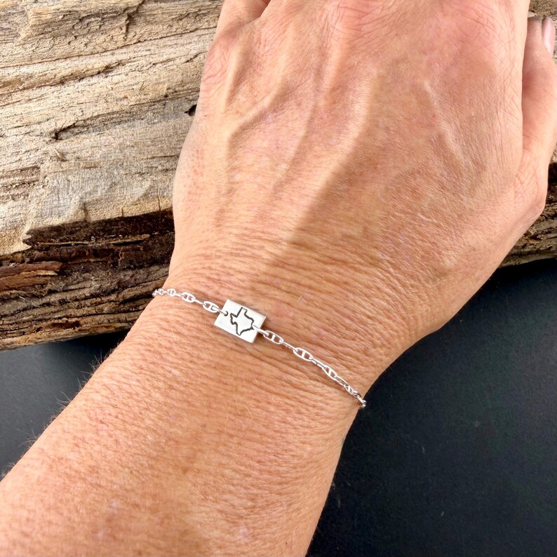May include: A silver bracelet featuring a rectangular charm with a cutout of the state of Texas. The bracelet has a delicate chain design and is displayed on a person's wrist.