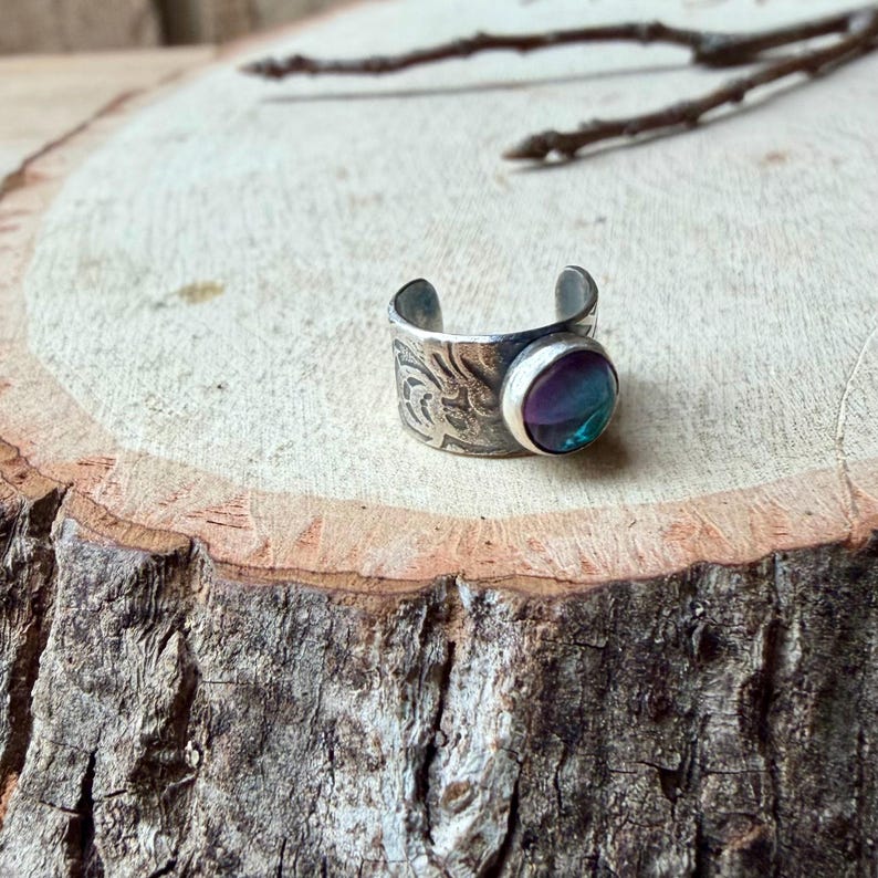 May include: A silver-toned adjustable ring with an engraved floral pattern. The ring features a round, iridescent stone with shades of purple, blue, and green. The ring is displayed on a wooden surface.