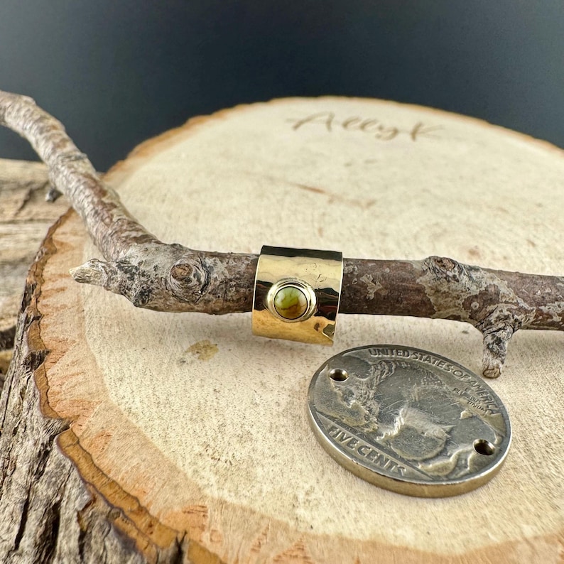 May include: A gold-toned ring with a hammered texture and a round, olive-green stone. The ring is displayed on a wooden branch, with a silver coin featuring a buffalo design nearby. The ring's design is simple and elegant.