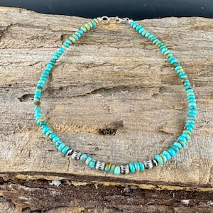 May include: Turquoise beaded necklace with silver accents. The necklace features small, round turquoise beads in shades of blue and green, interspersed with silver-colored, ribbed beads. The necklace is displayed on a wooden surface.