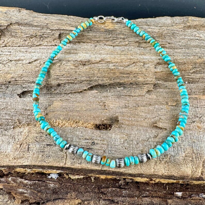 May include: Turquoise beaded necklace with silver accents. The necklace features small, round turquoise beads in shades of blue and green, interspersed with silver-colored, ribbed beads. The necklace is displayed on a wooden surface.