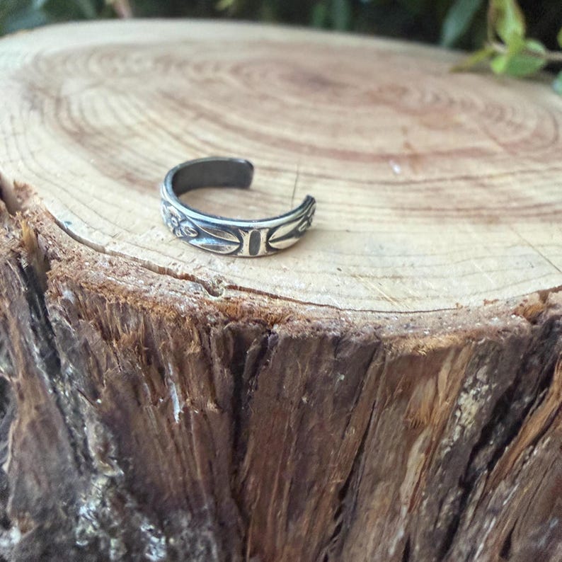 K&ouml;nnte beinhalten: Ein silberfarbener offener Ring mit einem sich wiederholenden gepr&auml;gten Blumen- und Blattmuster auf der Au&szlig;enfl&auml;che. Das Metall weist in den vertieften Bereichen des Designs ein dunkles, oxidiertes Finish auf, das die botanischen Details betont.