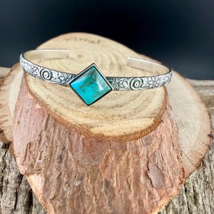 Puede incluir: Una pulsera de plata con una piedra preciosa de turquesa. La pulsera presenta un patr&oacute;n floral y de remolino a lo largo de la banda. La piedra de turquesa tiene forma cuadrada y est&aacute; engastada en un bisel de plata.