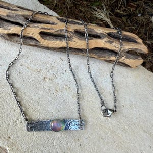 May include: A silver-toned necklace featuring a rectangular metal bar pendant with an embossed scroll pattern and a central oval iridescent cabochon stone. The pendant is attached to a delicate silver-toned chain with a lobster claw clasp.