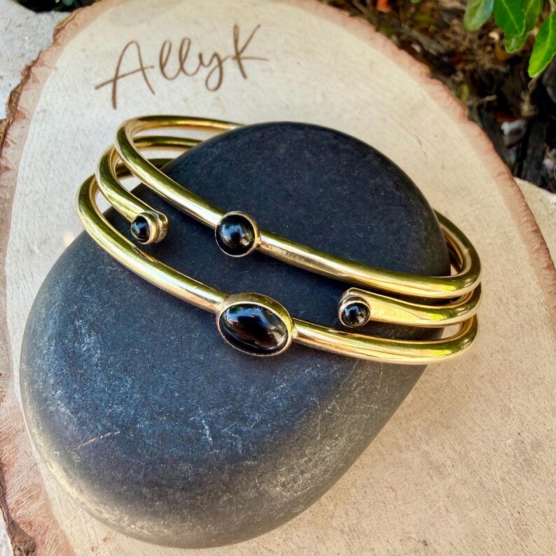 May include: Gold-toned bangles with black gemstone accents. The bracelets are stacked on a dark gray stone, with the word "Allyk" written in cursive on a wooden surface in the background. The jewelry is displayed in natural light.