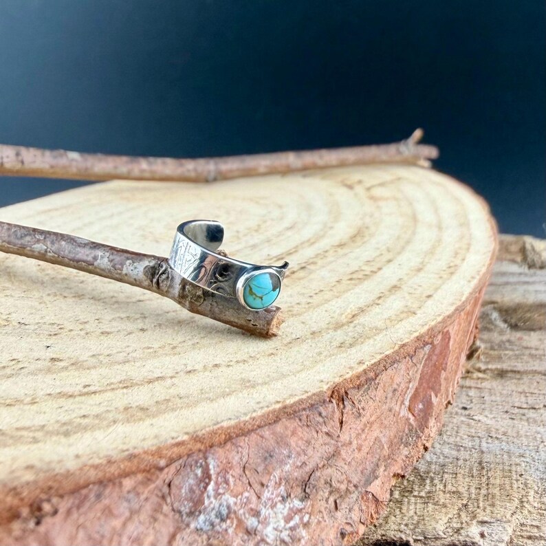 K&ouml;nnte beinhalten: Ein silberner Ring mit einem ovalen t&uuml;rkisfarbenen Stein. Der Ring hat ein offenes Design mit ge&auml;tzten Mustern auf dem Band. Der Ring wird auf einem Holzst&uuml;ck mit nat&uuml;rlichem, rustikalem Aussehen pr&auml;sentiert.