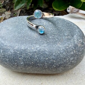 May include: A silver cuff bracelet featuring two round, blue-green gemstones. The bracelet has a curved design, with a gemstone at each end. The gemstones have an iridescent quality, reflecting light. The bracelet is displayed on a grey stone.
