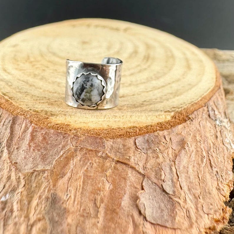 May include: A silver-toned ring with a wide band and a textured surface. The ring features a circular, light-colored stone with dark gray and black marbling. The ring is displayed on a wooden surface.
