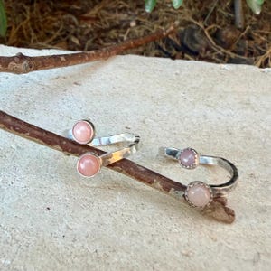 May include: Two open-style silver rings, each featuring two smooth, round, pale pink cabochon stones set at the ends of the band. The silver bands have a textured, hammered finish and are positioned resting on a thin, brown wooden twig.