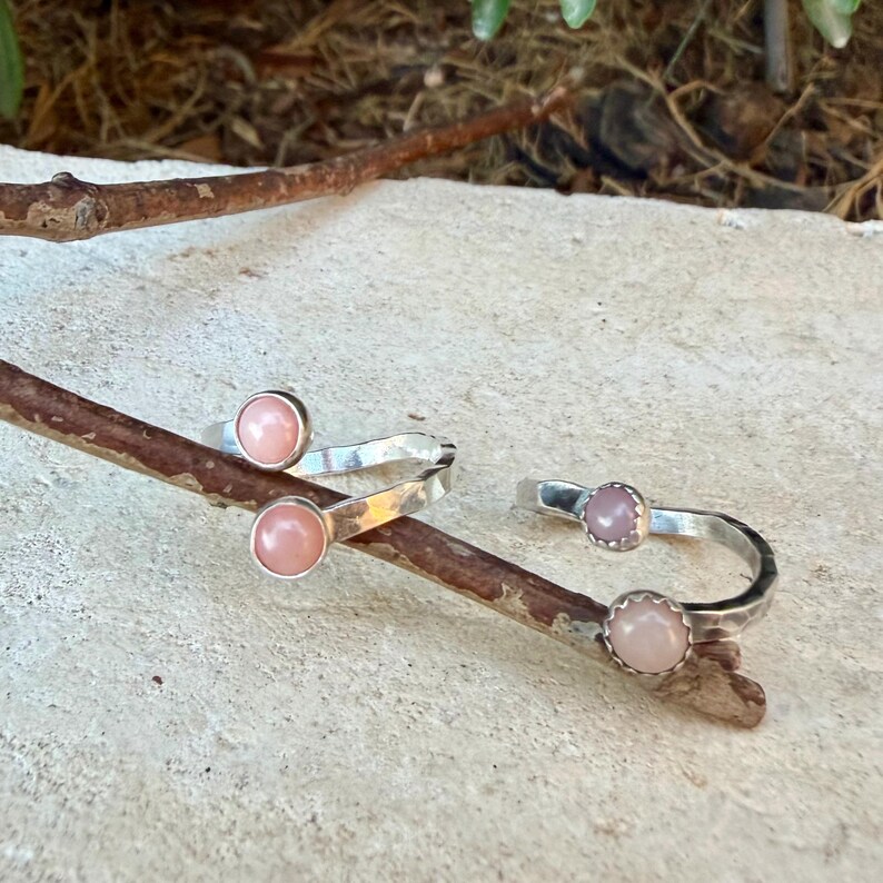 May include: Two open-style silver rings, each featuring two smooth, round, pale pink cabochon stones set at the ends of the band. The silver bands have a textured, hammered finish and are positioned resting on a thin, brown wooden twig.