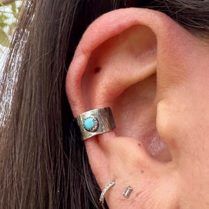 May include: A silver ear cuff with a textured surface and a round, light blue turquoise stone set in the center. The cuff is worn on the ear, with a small silver hoop earring and a stud earring visible below.