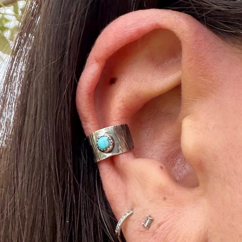 May include: A silver ear cuff with a textured surface and a round, light blue turquoise stone set in the center. The cuff is worn on the ear, with a small silver hoop earring and a stud earring visible below.
