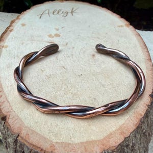 May include: A copper-colored cuff bracelet with a twisted design. The bracelet is open at the ends and rests on a wooden surface. The text "Allyk" is written on the wood.