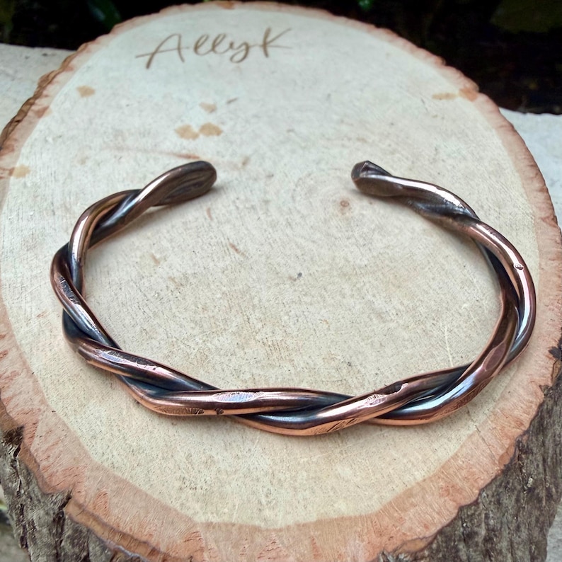 May include: A copper-colored cuff bracelet with a twisted design. The bracelet is open at the ends and rests on a wooden surface. The text "Allyk" is written on the wood.