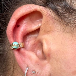 May include: Close-up of an ear adorned with a gold-colored ear cuff featuring a round, turquoise stone. The cuff has a textured design. Below, a small, diamond-studded earring and a small, rectangular stud are visible.