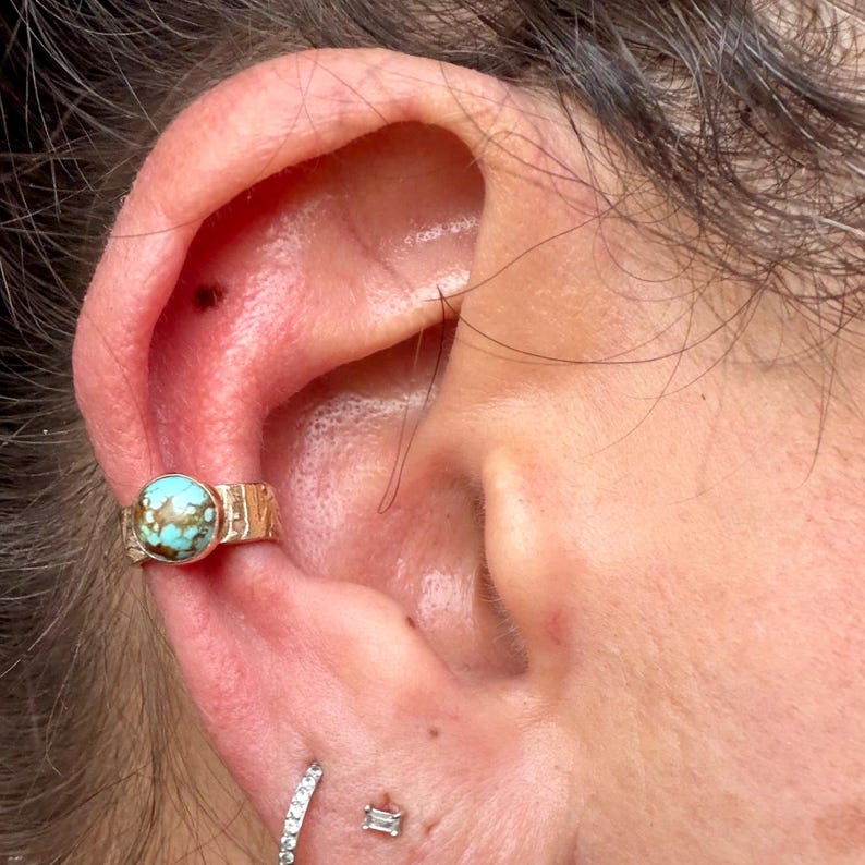 May include: Close-up of an ear adorned with a gold-colored ear cuff featuring a round, turquoise stone. The cuff has a textured design. Below, a small, diamond-studded earring and a small, rectangular stud are visible.