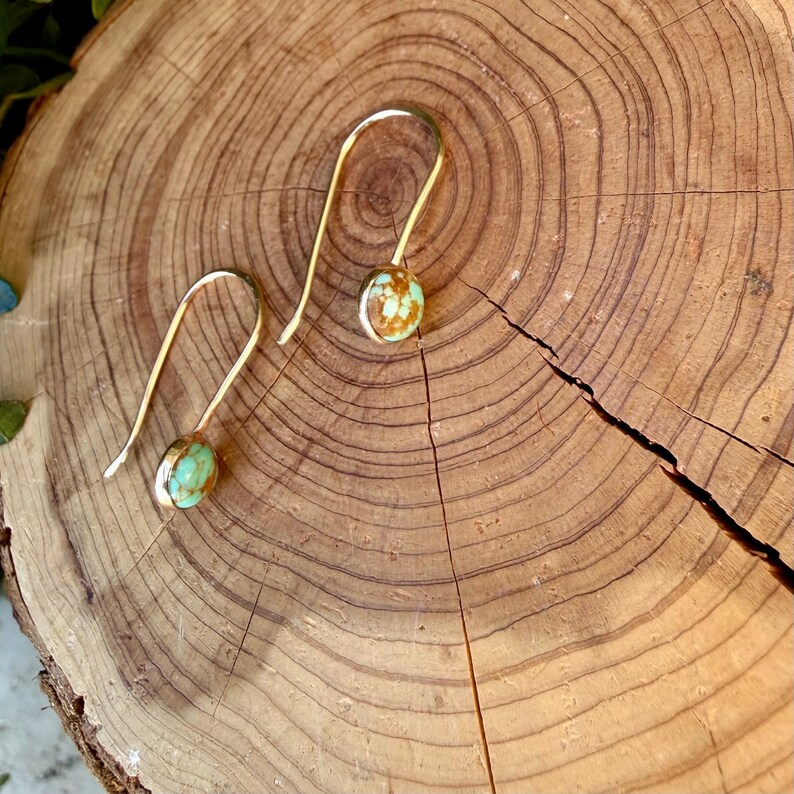 May include: A pair of gold-toned hook earrings featuring round, polished turquoise stones with brown matrix veining. The earrings have a simple, curved wire design that holds each stone in a secure setting, showcasing a smooth, rounded profile.